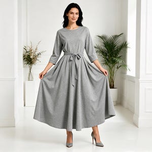 Puede incluir: Un vestido midi gris con cuello redondo y mangas tres cuartos. El vestido presenta un cinturón de tela a juego atado a la cintura y una falda acampanada. La modelo lleva tacones grises.