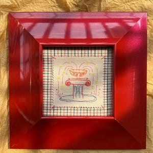 May include: A square, glossy red frame containing a piece of fabric with a cross-stitched fountain design. The fountain is embroidered in red, orange, and blue thread, set against a cream-colored fabric with a green and red plaid pattern background.