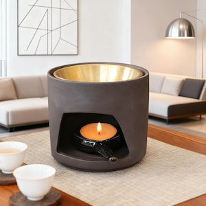 Puede incluir: Un difusor de aceites esenciales de cerámica marrón oscuro con interior dorado. Una vela encendida está dentro del difusor, y dos tazas de té blancas están en primer plano. El difusor está sobre una mesa de madera.