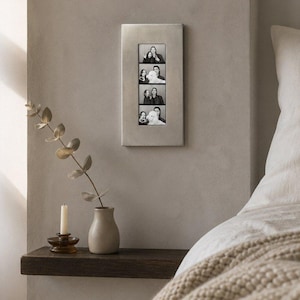 Puede incluir: Un marco de fotos rectangular de metal plateado montado en la pared, que contiene una tira vertical de cuatro fotografías en blanco y negro que muestran a dos personas y un perro en varias poses. El marco tiene un acabado liso y mate.