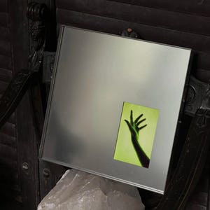 May include: A square, silver-toned metallic mirror featuring a rectangular inset panel. The inset displays a vibrant, lime-green background with a dark, silhouetted human hand reaching upward, fingers spread, creating a high-contrast visual effect.