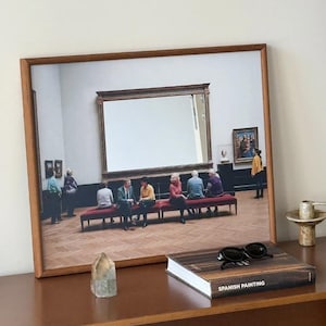 Puede incluir: Una impresión enmarcada que muestra el interior de un museo con personas sentadas en bancos rojos frente a un marco grande vacío, junto a un cristal, un libro titulado Spanish Painting y gafas de sol negras sobre una superficie marrón.