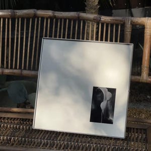 Puede incluir: Un espejo cuadrado con un marco plateado fino que presenta una pequeña fotografía rectangular en blanco y negro de una flor de cala colocada en la esquina inferior derecha. La superficie del espejo refleja el entorno y la luz.