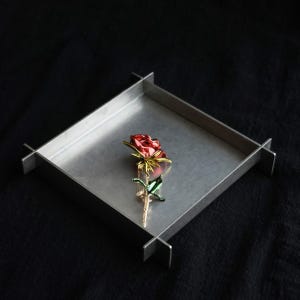 Puede incluir: Un broche con forma de rosa que presenta una cabeza de flor roja brillante con detalles en tono dorado y un tallo verde intenso con hojas, descansando dentro de una bandeja cuadrada de metal cepillado con bordes finos y elevados.