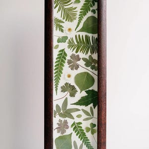 Puede incluir: Una pieza de arte mural alta y rectangular con un marco de madera marrón oscuro que encierra un fondo blanco. La composición muestra una variedad de hojas verdes prensadas, helechos y pequeñas flores blancas con centros amarillos.