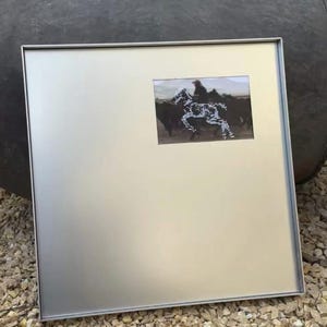 Pode incluir: Uma moldura metálica quadrada de tom prateado com um acabamento liso e mate. Uma fotografia retangular está inserida no quadrante superior direito, retratando um cavaleiro a cavalo com a silhueta pontilhada e clara de um cavalo.