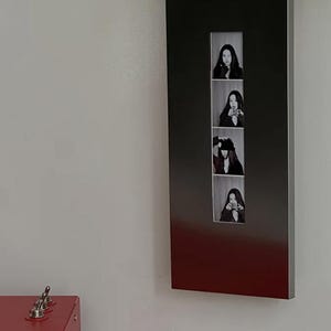 Puede incluir: Un marco de pared alto, rectangular y de color oscuro que presenta una tira vertical de cuatro fotografías en blanco y negro. Cada foto muestra a una persona en diferentes poses. Una parte de un objeto rojo con interruptores metálicos es visible en la esquina.