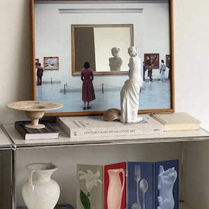 Puede incluir: Una lámina enmarcada de un interior de museo con una persona y una estatua, un cuenco beige texturizado con pie, un libro titulado Contemporary Installation Art, una estatua blanca, un jarrón blanco moteado y un biombo de cuatro paneles.