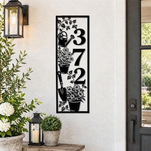 May include: A vertical rectangular black metal house number sign featuring the digits 372. The design includes cutouts of a watering can, two potted flower arrangements, a hand trowel, and trailing vines against a white background.