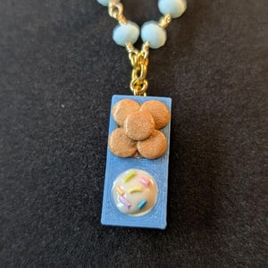May include: A necklace featuring a blue pendant with a miniature cookie design. The pendant has a tray of cookies and a frosted cookie with sprinkles. The necklace has light blue beads and gold-colored chain.