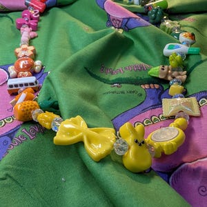 Könnte beinhalten: Eine farbenfrohe Perlenkette mit verschiedenen Anhängern, darunter eine gelbe Schleife, ein Hase und ein Bus. Die Halskette hat rosa, orange, gelbe und klare Perlen. Die Halskette wird auf einem grünen Stoff mit einem Cartoon-Design präsentiert.
