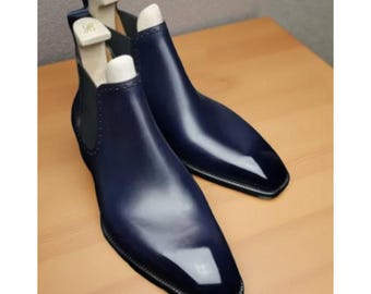 May include: A pair of navy blue leather Chelsea boots with a high-gloss finish. The boots have a square toe and elastic side panels. Wooden shoe trees are inserted inside the boots to maintain their shape.