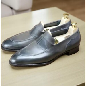 May include: A pair of gray leather slip-on dress shoes with wooden shoe trees inside. The shoes have a rounded toe and a low heel. The leather has a slight sheen, and the shoes are displayed on a light-colored wooden surface.