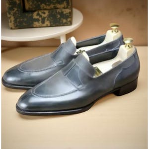 May include: A pair of gray leather slip-on loafers with a rounded toe and a low heel. The shoes are displayed with wooden shoe trees inside. The leather has a slight sheen, and the shoes appear to be new.