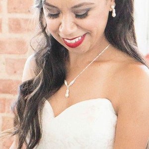May include: A strapless white gown featuring a sweetheart neckline, paired with a delicate silver necklace with a teardrop pendant and matching teardrop earrings. The jewelry set is crafted with small clear stones and a white pearl-like drop.