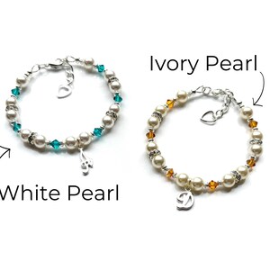 May include: Two pearl bracelets with charms. The bracelet on the left has white pearls and teal blue crystals with a music note charm. The bracelet on the right has ivory pearls and amber crystals with a letter D charm.  White Pearl and Ivory Pearl are written below the bracelets.