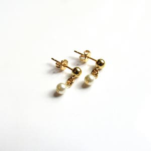 Puede incluir: Un par de pendientes dorados con una peque&ntilde;a perla blanca colgante. Los pendientes tienen un dise&ntilde;o sencillo, con un perno redondo dorado y una delicada cadena que sujeta la perla. Fondo blanco.