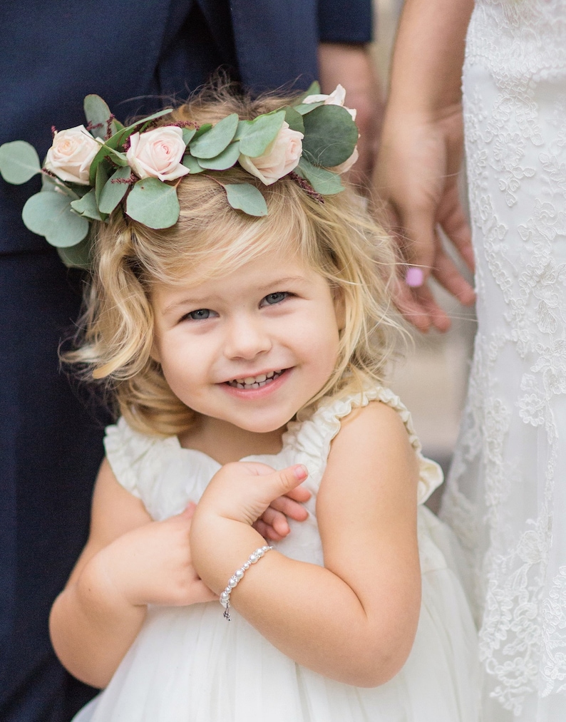 May include: A young girl wearing a white dress and a flower crown with pink roses and green leaves. She is smiling and holding her hands together, wearing a pearl bracelet on her wrist.