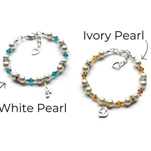 May include: Two pearl bracelets with charms. The bracelet on the left has white pearls and teal blue crystals with a music note charm. The bracelet on the right has ivory pearls and amber crystals with a letter D charm.  White Pearl and Ivory Pearl are written below the bracelets.