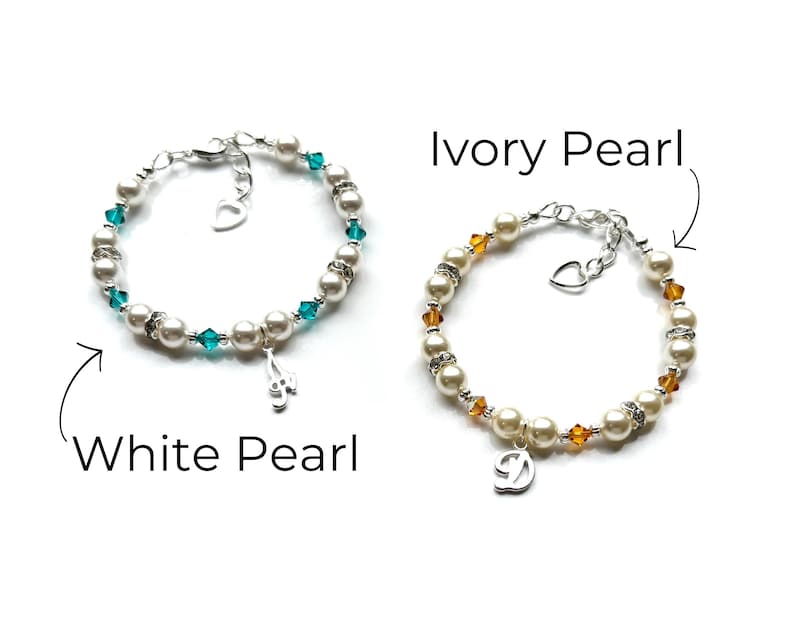 May include: Two pearl bracelets with charms. The bracelet on the left has white pearls and teal blue crystals with a music note charm. The bracelet on the right has ivory pearls and amber crystals with a letter D charm.  White Pearl and Ivory Pearl are written below the bracelets.