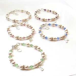 May include: Four pearl bracelets with crystal accents and silver charms. Each bracelet has a different colored crystal: purple, blue, green, and clear. The charms are silver hearts and letters.
