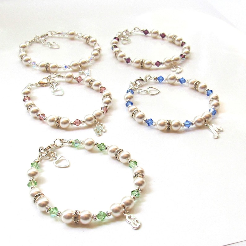 May include: Four pearl bracelets with crystal accents and silver charms. Each bracelet has a different colored crystal: purple, blue, green, and clear. The charms are silver hearts and letters.