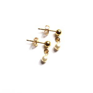 Puede incluir: Un par de pendientes dorados con una peque&ntilde;a perla blanca colgante. Los pendientes tienen un dise&ntilde;o de poste y bola, con una peque&ntilde;a perla suspendida de la bola. Los pendientes est&aacute;n sobre un fondo blanco.