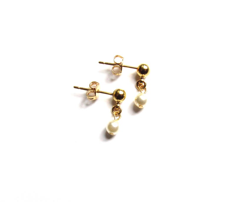 Puede incluir: Un par de pendientes dorados con una peque&ntilde;a perla blanca colgante. Los pendientes tienen un dise&ntilde;o de poste y bola, con una peque&ntilde;a perla suspendida de la bola. Los pendientes est&aacute;n sobre un fondo blanco.