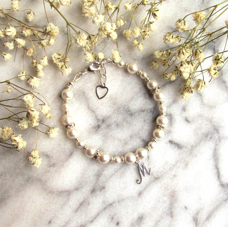 May include: A silver bracelet with white pearl beads, a small silver heart charm, and a silver letter 'M' charm. The bracelet is on a white marble surface with white flowers.