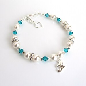 May include: A white pearl and teal blue crystal bracelet with a silver charm. The charm is a stylized letter J.