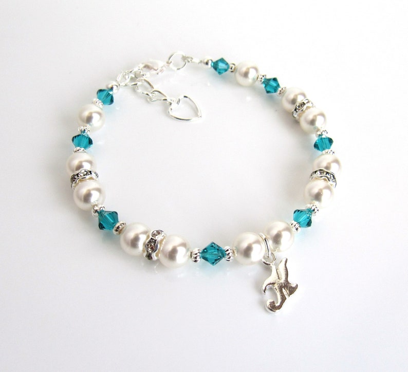 May include: A white pearl and teal blue crystal bracelet with a silver charm. The charm is a stylized letter J.