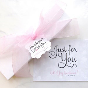 May include: A white gift box with a pink ribbon and a tag that says "Handmade Just for You Mel Joy Creations Jewelry". The box is sitting on a white surface with a card that says "Just for You Mel Joy Creations Jewelry".