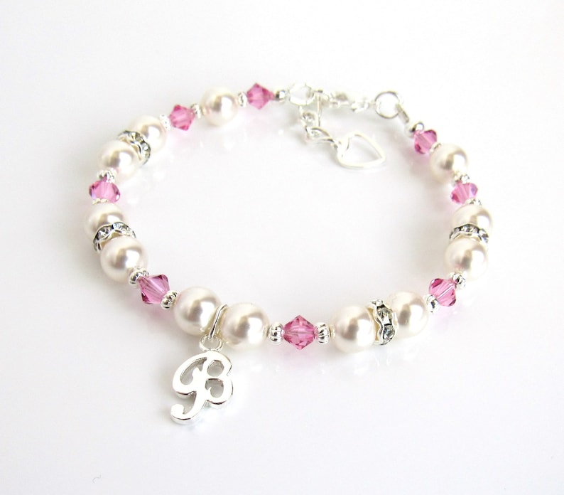 May include: A silver bracelet with pink crystals and white pearls. The bracelet has a silver charm with the letter "B" on it.