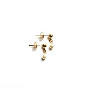 Puede incluir: Un par de pendientes dorados con una peque&ntilde;a perla blanca colgante. Los pendientes tienen un dise&ntilde;o cl&aacute;sico, con un perno redondo y una delicada gota de perla. Se presentan sobre un fondo blanco.