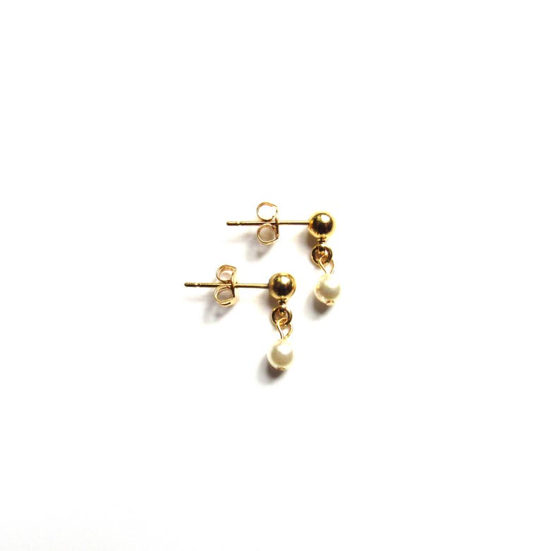 Puede incluir: Un par de pendientes dorados con una peque&ntilde;a perla blanca colgante. Los pendientes tienen un dise&ntilde;o cl&aacute;sico, con un perno redondo y una delicada gota de perla. Se presentan sobre un fondo blanco.