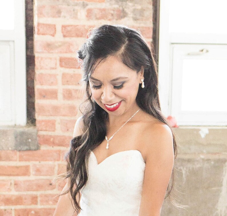 May include: A strapless white gown featuring a sweetheart neckline, paired with a delicate silver necklace with a teardrop pendant and matching teardrop earrings. The jewelry set is crafted with small clear stones and a white pearl-like drop.