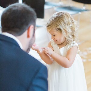 May include: A young girl in a white dress is looking down at a silver bracelet on her wrist. She is standing in front of a man in a dark suit.