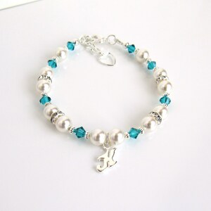 May include: A silver bracelet with white pearls and teal blue crystals. The bracelet has a silver charm with the letter "K" on it.