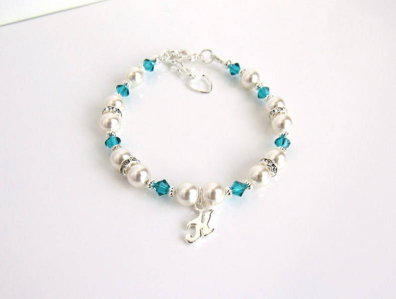 May include: A silver bracelet with white pearls and teal blue crystals. The bracelet has a silver charm with the letter "K" on it.