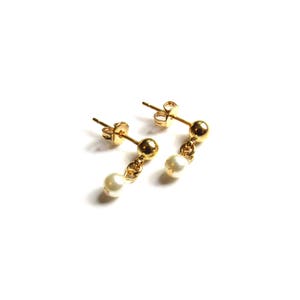 Puede incluir: Un par de pendientes dorados con una peque&ntilde;a perla blanca colgante. Los pendientes tienen un dise&ntilde;o de poste y bola, con una peque&ntilde;a perla suspendida de una delicada cadena. Los pendientes est&aacute;n sobre un fondo blanco.