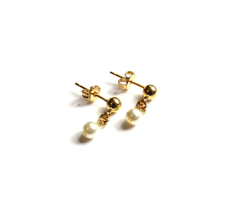 Puede incluir: Un par de pendientes dorados con una peque&ntilde;a perla blanca colgante. Los pendientes tienen un dise&ntilde;o de poste y bola, con una peque&ntilde;a perla suspendida de una delicada cadena. Los pendientes est&aacute;n sobre un fondo blanco.