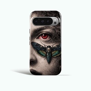 May include: A phone case featuring a detailed illustration of a face with a red eye and a Death's-head Hawkmoth. The moth has a skull design on its body and is positioned over the eye. The case has a black and silver camera module.