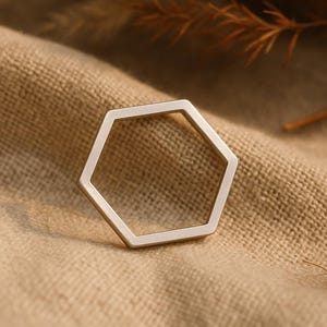 Puede incluir: Un anillo hexagonal geométrico de color plateado. El anillo se encuentra sobre un fondo de tela texturizada de tonos neutros, con follaje seco en la parte superior de la imagen. El diseño del anillo es simple y moderno.