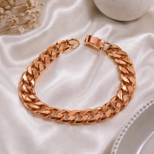 May include: A rose gold curb chain bracelet. The bracelet features a series of interlocking, rounded links. The clasp is a matching rose gold tone. The bracelet is displayed on a white, satin-like surface.