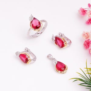 Puede incluir: Un conjunto de joyas con un anillo, pendientes y un colgante. Cada pieza tiene una piedra preciosa roja rubí en forma de lágrima engastada en metal plateado y dorado. El anillo y los pendientes tienen un pequeño acento de piedra transparente.