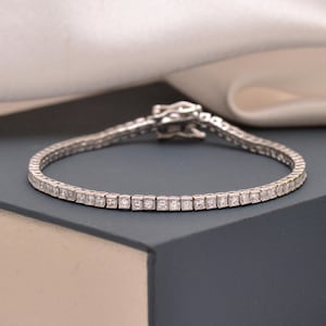 Op de afbeelding: Een zilveren tennisarmband met vierkant geslepen edelstenen. De armband ligt op een donkerblauwe ondergrond, met witte satijnstof op de achtergrond. De sluiting van de armband is zichtbaar.