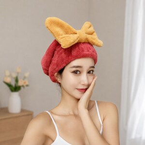 Pode incluir: Uma toalha de cabelo vermelha em tecido felpudo texturizado, com um laço amarelo grande e macio no topo. O acessório foi concebido para prender o cabelo, com o laço a proporcionar um elemento decorativo na parte frontal da touca.