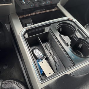 May include: Black car console organizer with a divided storage compartment, cup holders, and a USB port. The organizer holds a car key, glasses, and other small items. The interior is black, and the surrounding trim is silver.