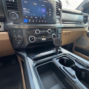 May include: Interior view of a vehicle's dashboard, featuring a large infotainment screen displaying radio information. The dashboard is primarily black and tan, with various controls and a gear shift lever. Cup holders and storage compartments are visible.