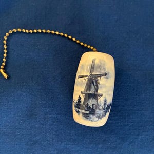May include: A ceramic light pull featuring a blue and white windmill scene. The pull has a rectangular shape with rounded edges and is attached to a gold-colored beaded chain. The background is a solid blue.